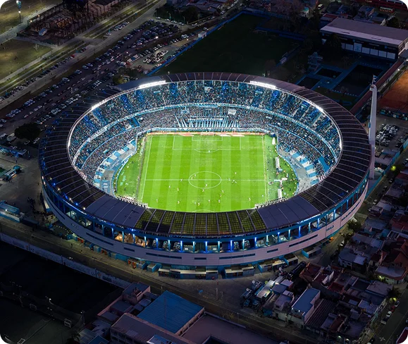 Vue aérienne d'un stade bien éclairé et bondé la nuit pendant un match de football. Le terrain vert est entouré de fans en train d'applaudir, créant une atmosphère vivante.